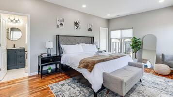 A modern bedroom with a large bed, bedside tables, a grey sofa, a round ottoman, and a potted plant.