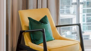 Un fauteuil jaune avec un coussin vert, dans une pièce dotée d’une grande fenêtre donnant sur un immeuble.