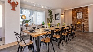 A dining area with a long wooden table set for six, surrounded by black chairs. There are potted plants and a large window with blinds. - Maison Rioma - Grande maison à 200m de la plage (Plounéour-Trez)