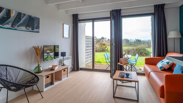 Una sala de estar moderna con un sofá, una silla, una mesa de centro y un televisor. Hay una puerta corrediza de vidrio que da a un balcón con mobiliario exterior.