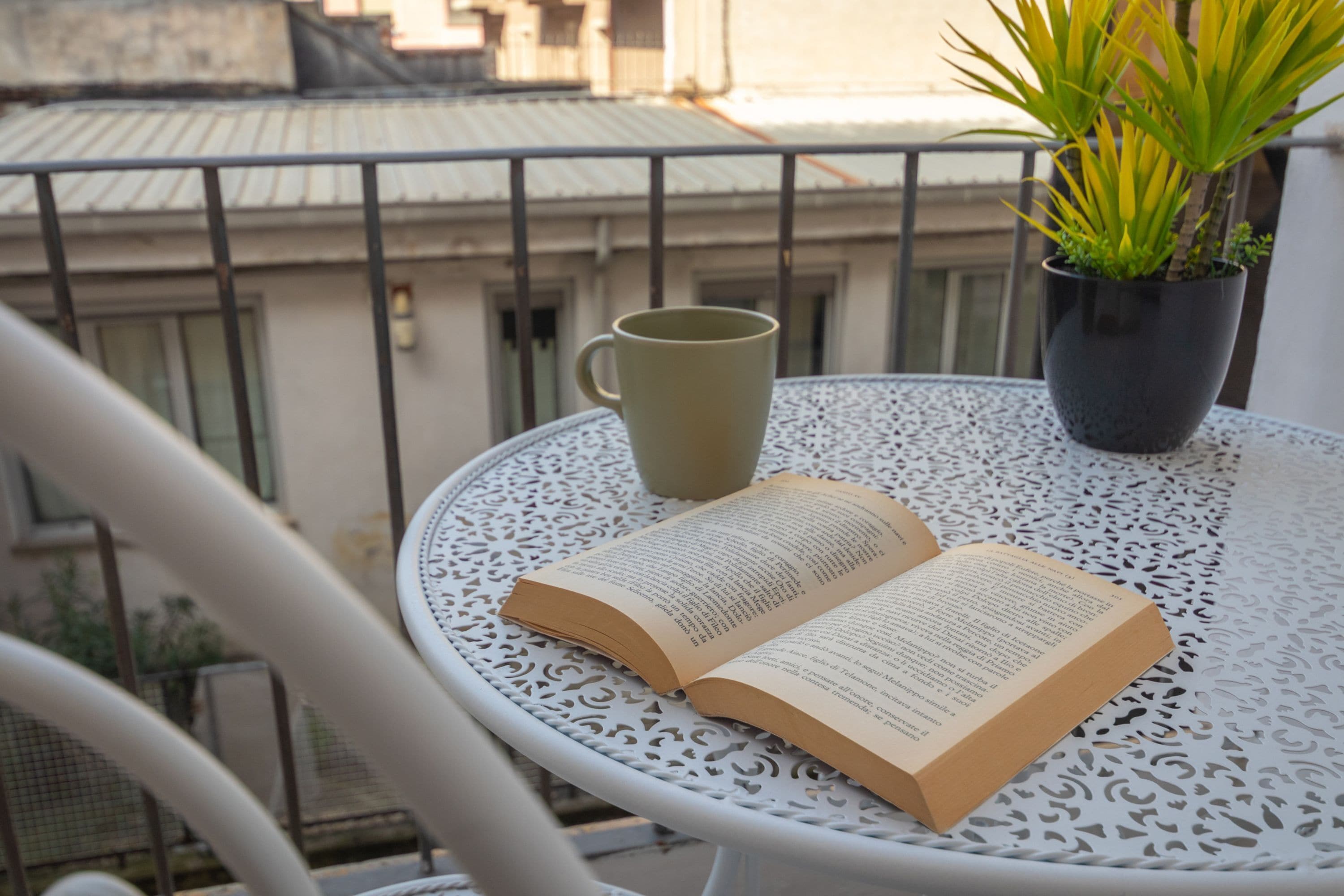 An open book on a white metal table with a green mug and a potted plant on a balcony.