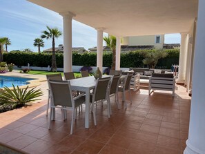 A covered outdoor patio with white furniture, a pool, and a view of the surrounding greenery and buildings.