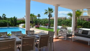 A covered outdoor dining area with a pool view, white furniture, and lush greenery. - Beautiful Villa Sunflower with private pool and garden, nearby the beaches. (Xabia)