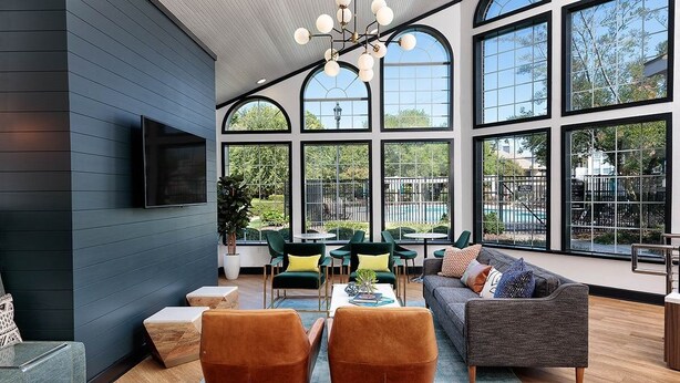 A modern living room with a flat-screen TV mounted on the wall, a grey sofa, two brown armchairs, a coffee table, and a large window with a view of the outdoors.