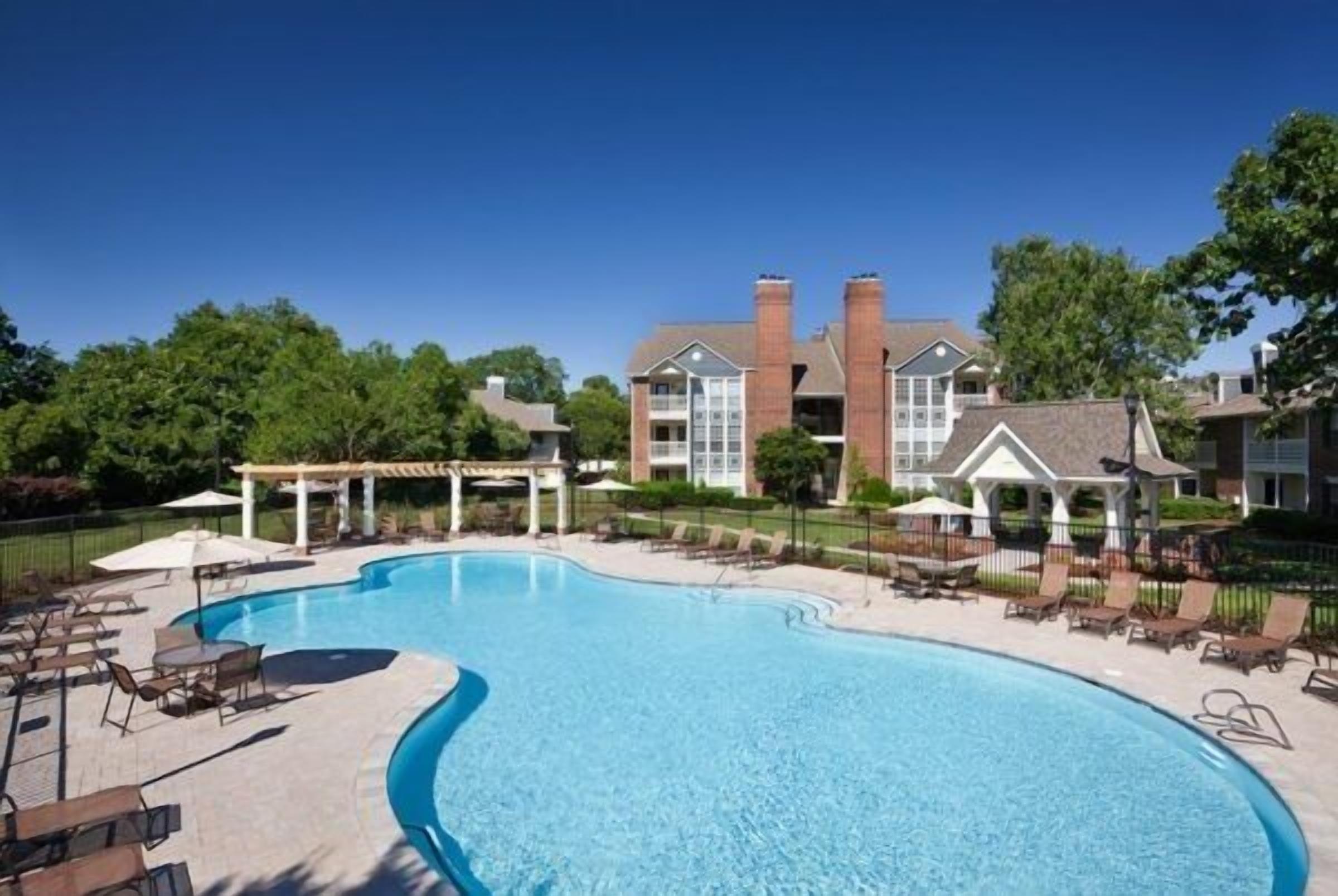 A swimming pool with lounge chairs and umbrellas, surrounded by a building with brick chimneys and a covered patio area.
