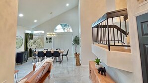 Amplia sala de estar con techo alto, una gran lámpara de araña, comedor con mesa redonda y un banco de madera.