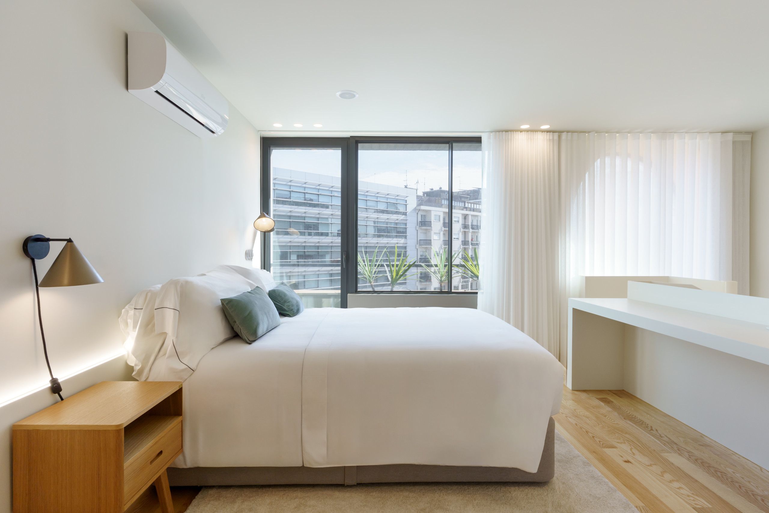 A modern bedroom with a large bed, bedside table, and a view of buildings through the window.