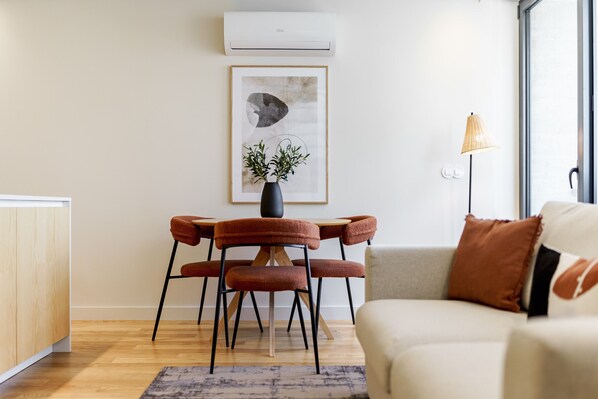 A modern living room with a dining area, featuring a sofa, a table with chairs, a vase with plants, and a framed artwork on the wall.
