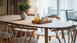 Una mesa de comedor de madera con una bandeja de alimentos para el desayuno, incluyendo un vaso de jugo de naranja, un frasco de mermelada y un tazón pequeño de cereal. Hay sillas de madera alrededor de la mesa, y una planta en maceta está sobre la mesa.