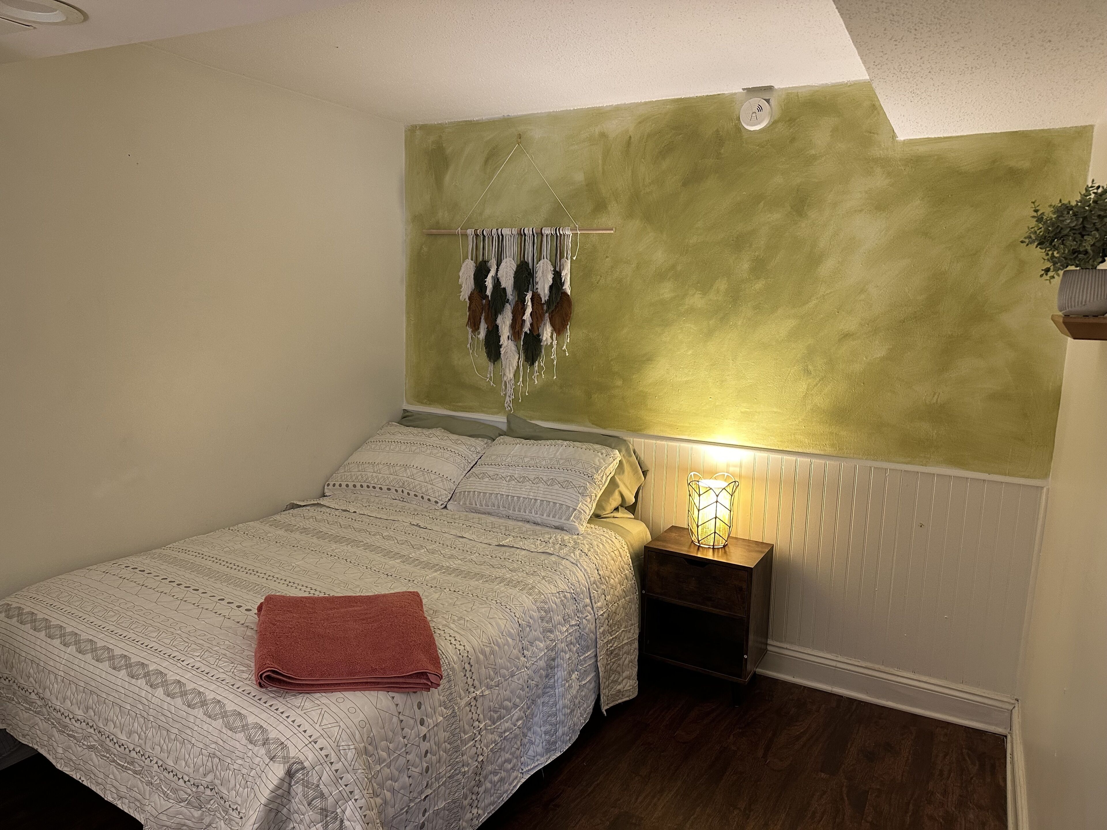 Ein Schlafzimmer mit einem Bett, einem Nachttisch, einer Wandlampe und einem hängenden Wandbehang aus Makramee.