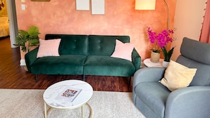 A living room with a green sofa, a grey armchair, a small white table with a magazine, a floor lamp, and a potted plant.