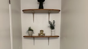A hallway with white walls and dark wooden flooring. There are two wooden shelves mounted on the wall, each holding a potted plant and a decorative item.