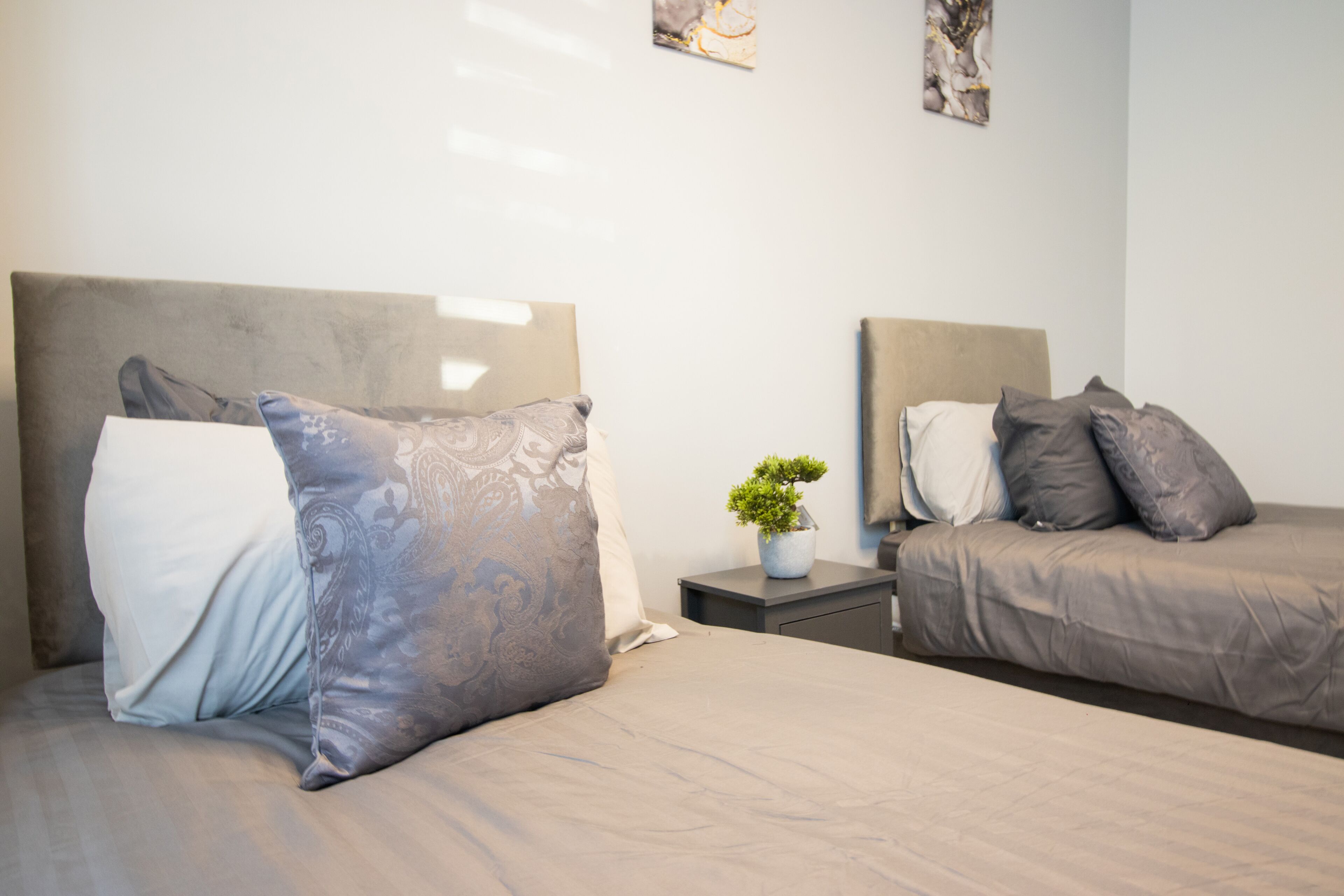 A hotel room with two beds, each with a headboard, a bedside table with a plant, and artwork on the wall.