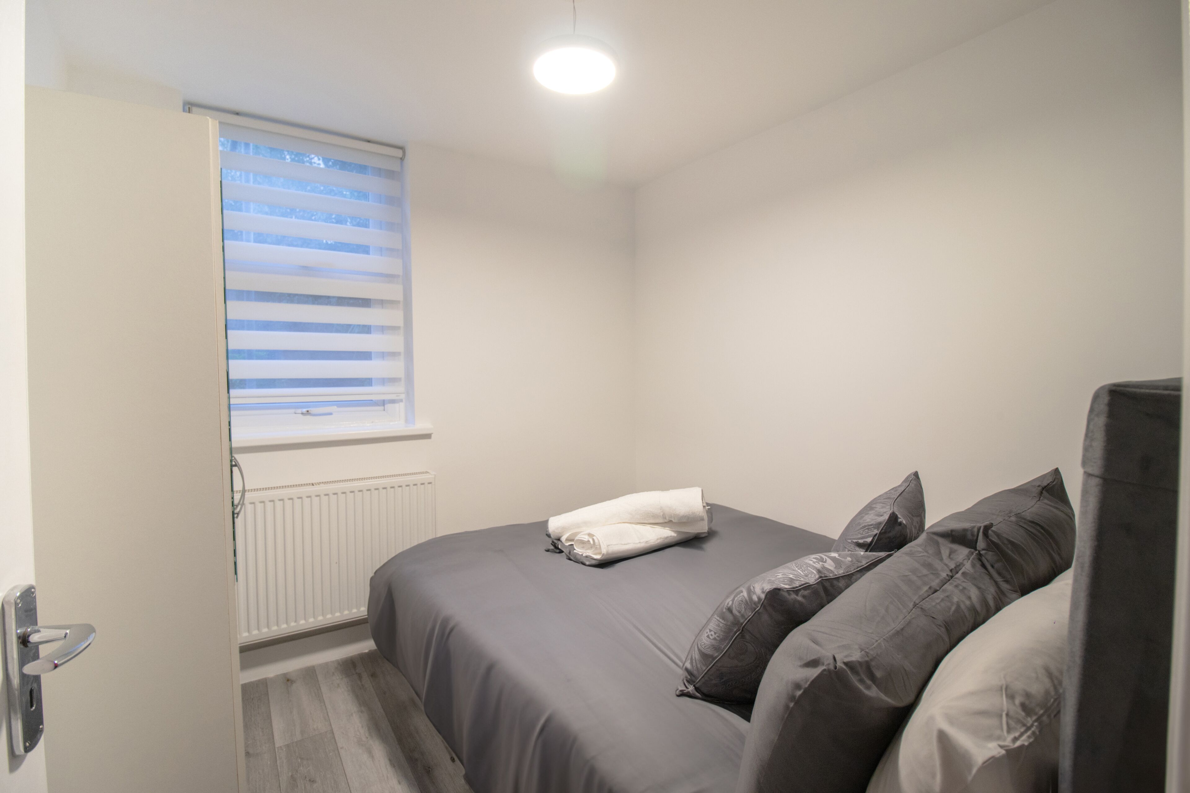 Une petite chambre à coucher, bien rangée, avec un lit, des oreillers, un radiateur et une fenêtre équipée de stores.