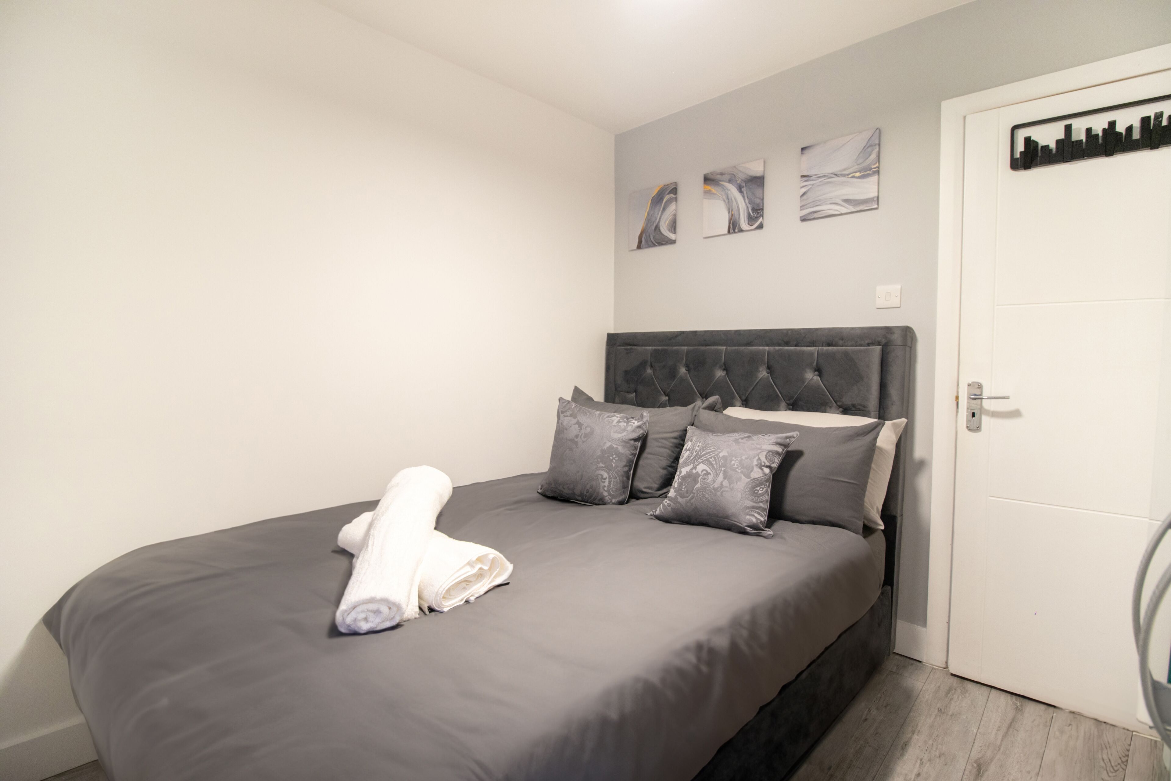 A neatly made bed with grey bedding and a rolled white towel, a grey upholstered headboard, and two abstract wall art pieces.