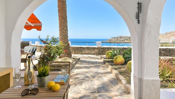 A stone-paved patio with a view of the sea, a table with a bottle, glasses, lemons, and a book.