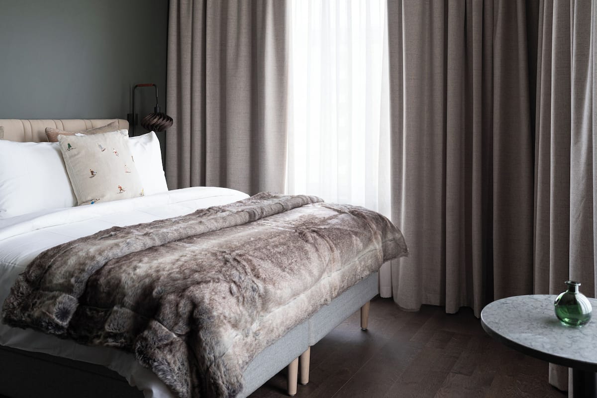 A bedroom with a bed, pillows, a fur throw, a bedside lamp, a window with curtains, and a small round table with a green bottle.