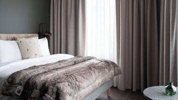 Quarto com cama, almofadas, manta de pele, abajur, janela com cortinas e uma pequena mesa redonda com uma garrafa verde.