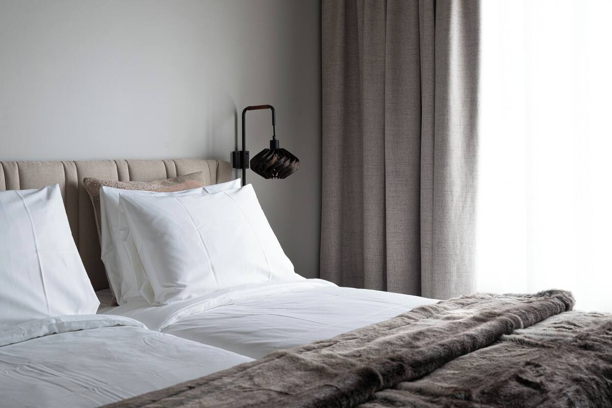 A neatly made bed with white linens and a grey blanket, a headboard, and a wall-mounted lamp.