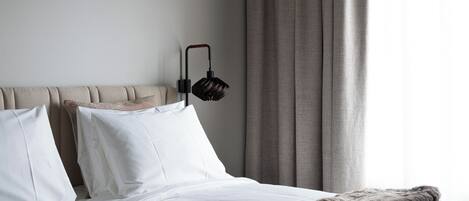 Una cama bien tendida con sábanas blancas y una manta gris, cabecero y una lámpara fijada a la pared.