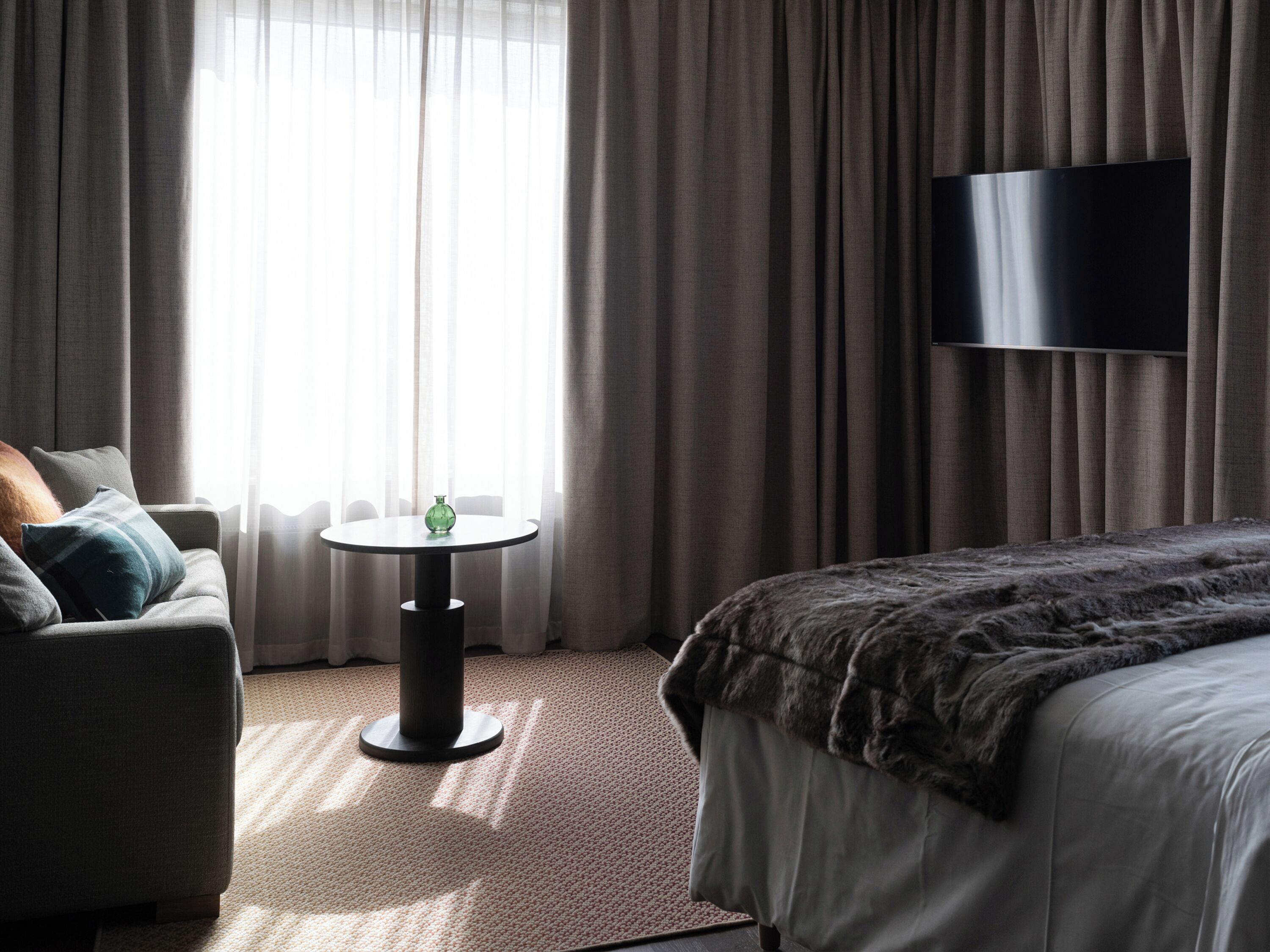 A hotel room with a bed, a sofa, a small round table with a green vase, and a large flat-screen TV on the wall.