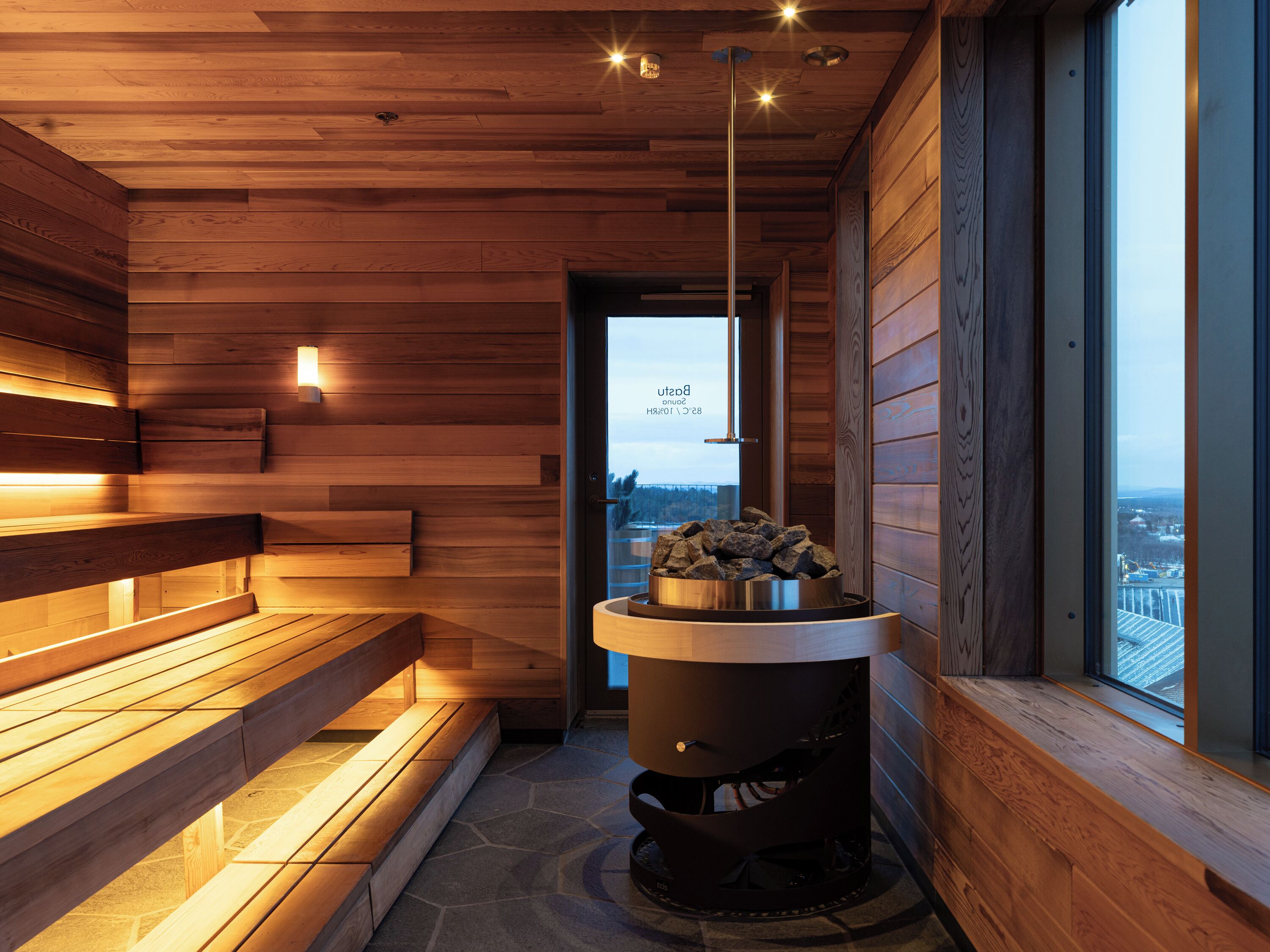 A wooden interior with benches and a stone floor, a fireplace, and a view through a door.