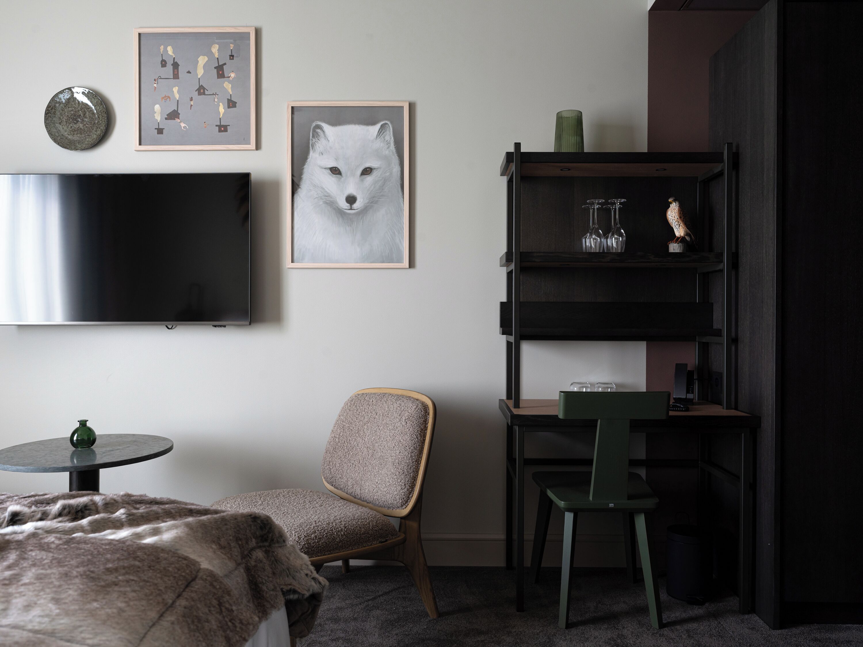 A modern room with a large flat-screen TV, a framed fox artwork, a small round table with a green bottle, two chairs, and a dark wooden bookshelf with decorative items.