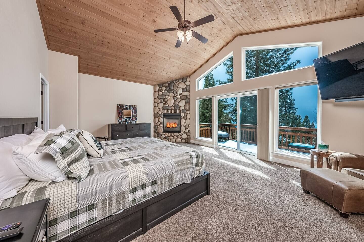 Une chambre à coucher comprenant un lit, une cheminée en pierre, un ventilateur de plafond et de grandes fenêtres donnant sur l’extérieur.