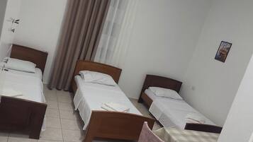 Una habitación de hotel con tres camas individuales, una silla de madera, una mesita y una cortina.