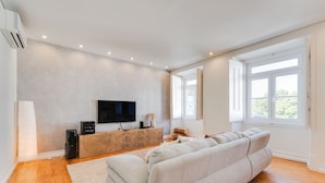 A modern living room with a large beige sofa, a flat-screen TV, a wooden coffee table, and a large window providing natural light. - Charming Graça Apartment, Lisbon City Centre, TTL354a (Lisboa ciudad)