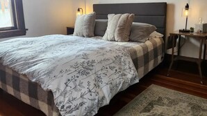 A bedroom with a large bed, a bedside table with a lamp, a wall-mounted shelf with decorative items, and a window with curtains. - Historic Craftsman 3 Bedroom Home in Uptown Butte (Butte)
