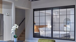 A modern hotel room with a large sliding glass door leading to a bedroom, a small table with a plant, a green chair, and a staircase. - Riversuites at The Battery (Philadelphia)