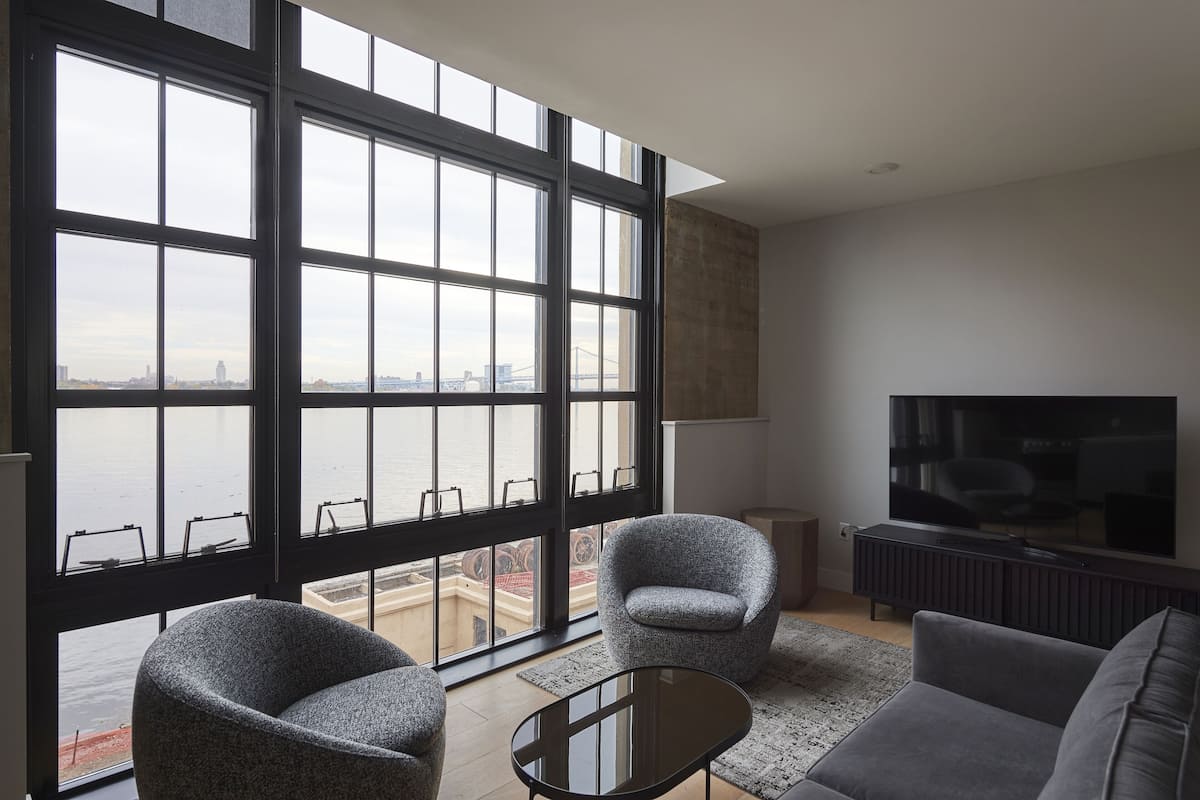 A modern living room with large windows, a flat-screen TV, and two armchairs.