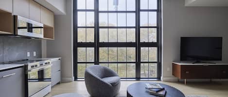 A modern kitchen with a large window, a grey armchair, a round coffee table, and a flat-screen TV.