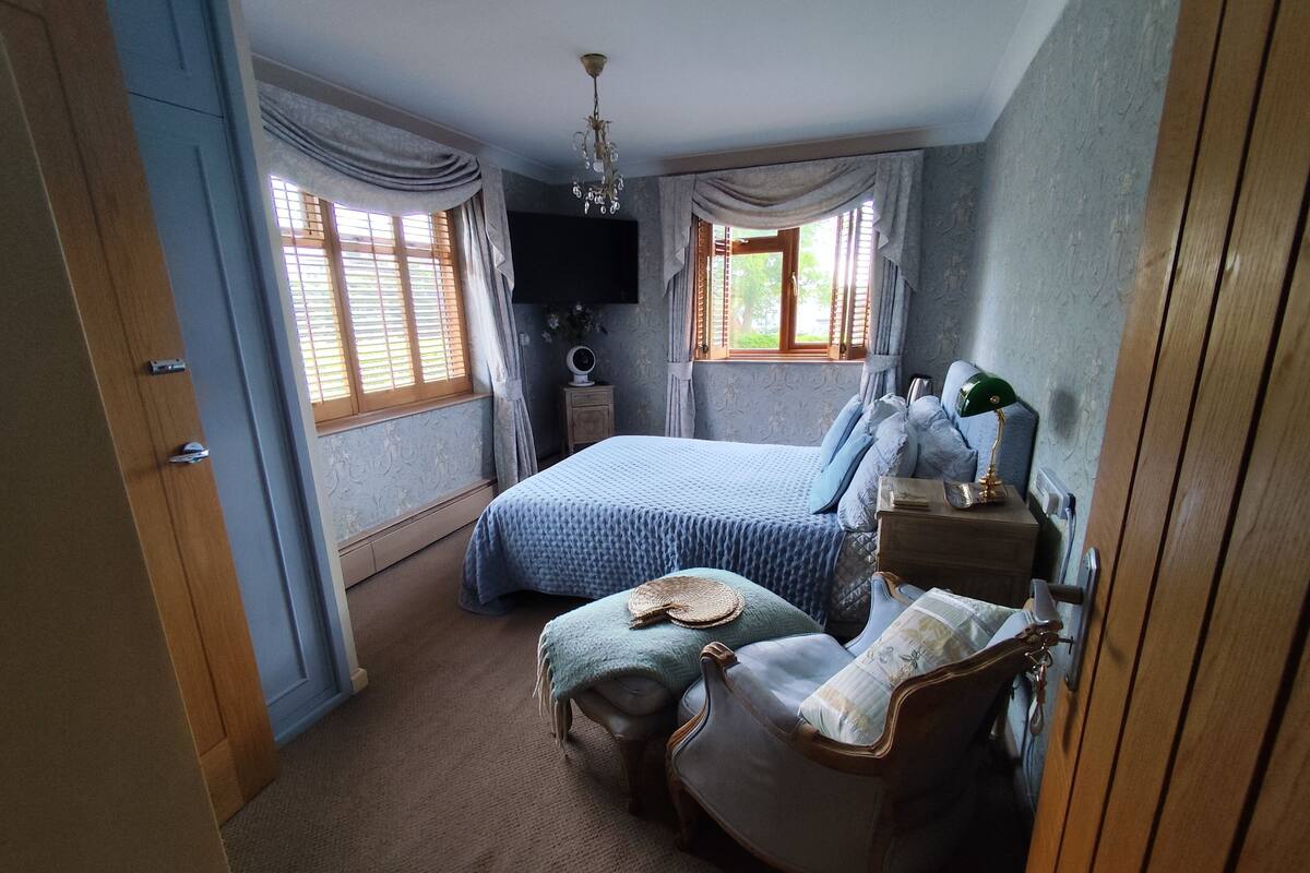 Een slaapkamer met een bed, een stoel, een nachtkastje, een televisie en een raam met gordijnen.