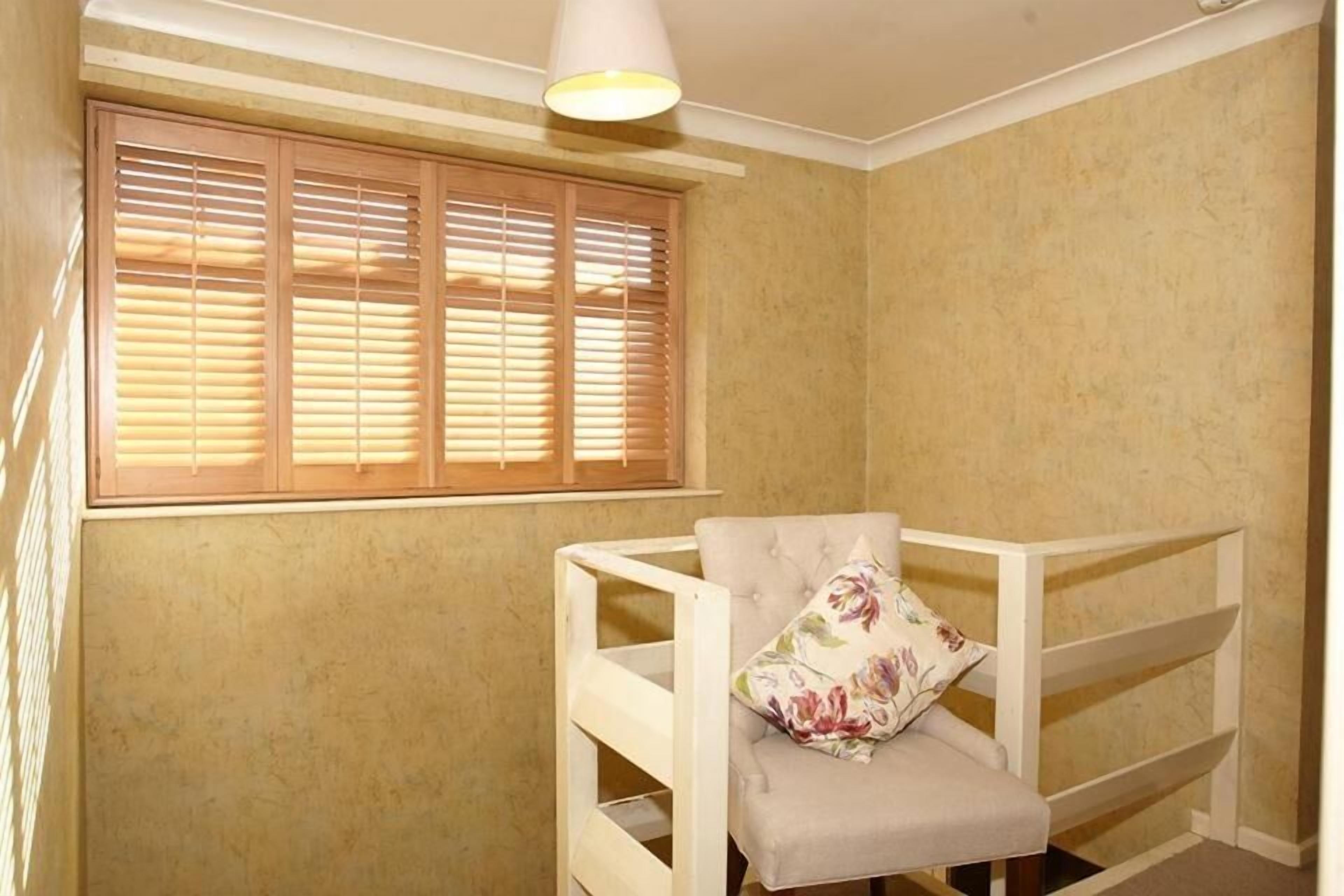 A room with a window with wooden blinds, a white upholstered chair with a floral pillow, and a white shelf unit.