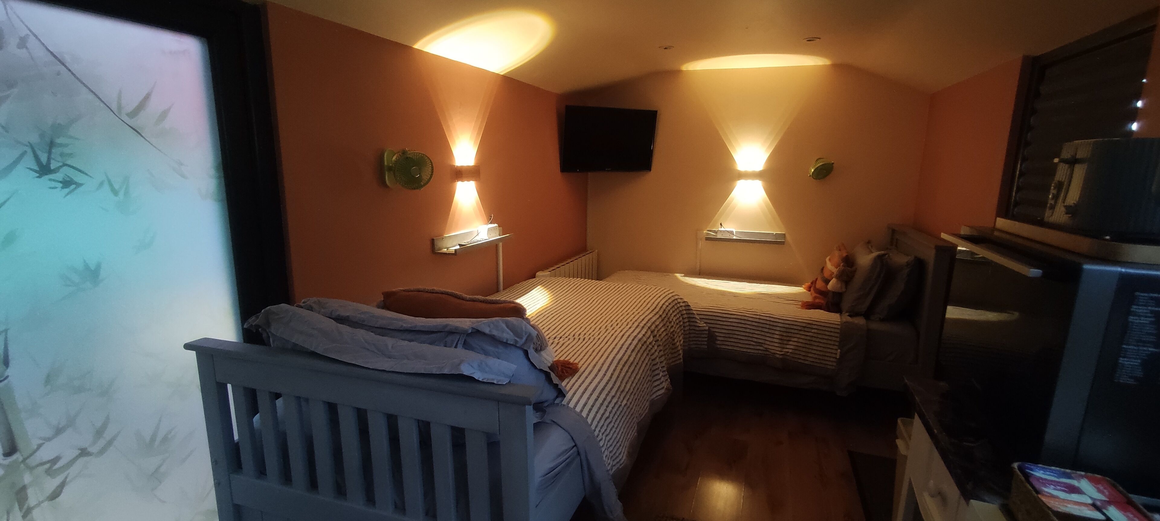 Une chambre de taille modeste, équipée de deux lits, d’une petite kitchenette et d’un téléviseur fixé au mur.