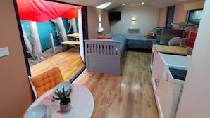 Een compacte kamer met een bed, een klein tafeltje met stoel, een kitchenette en uitzicht op een buitengebied met een rode luifel.