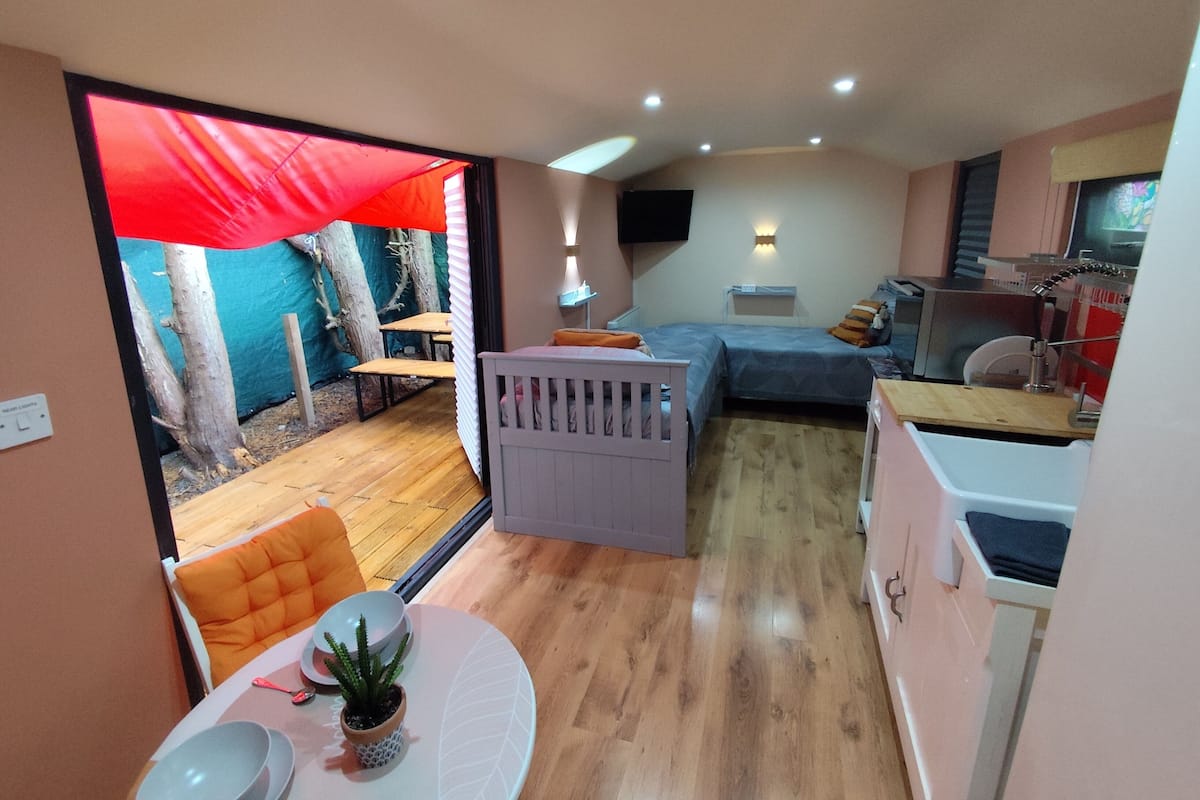 Een compacte kamer met een bed, een klein tafeltje met stoel, een kitchenette en uitzicht op een buitengebied met een rode luifel.