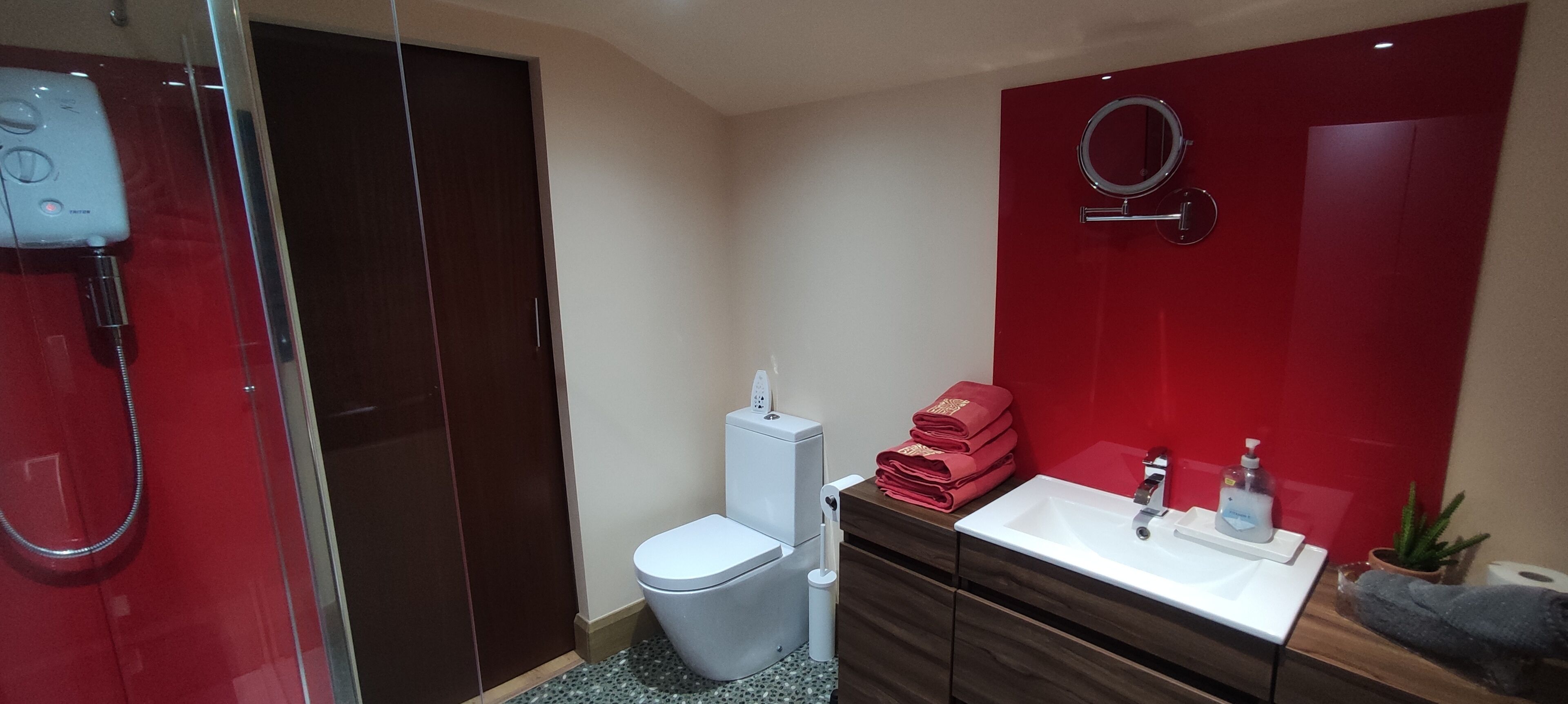 A modern bathroom with a glass shower enclosure, a white toilet, a vanity with a sink, and a red accent wall.