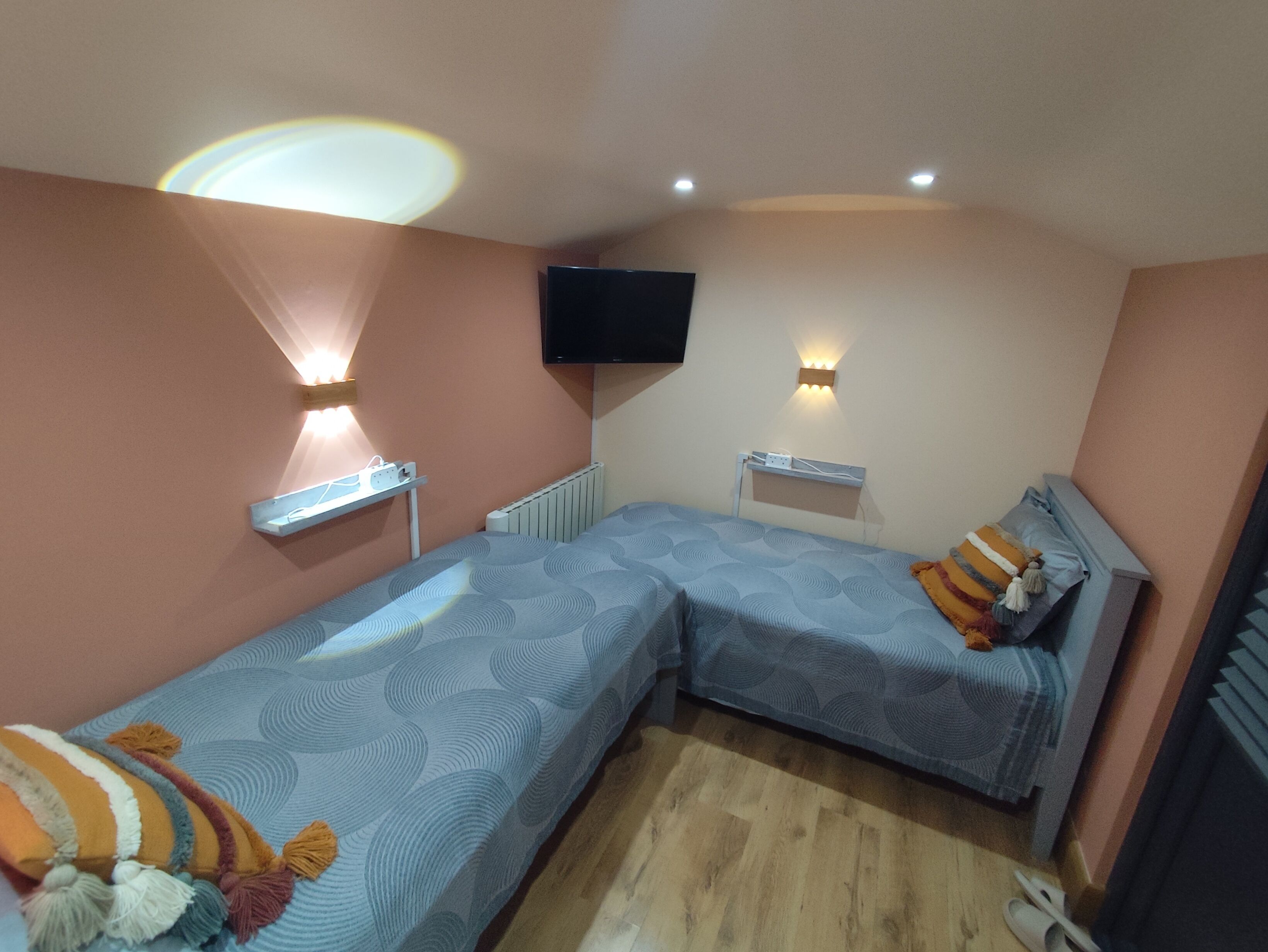 A room with two beds, a TV mounted on the wall, and a ceiling light fixture.