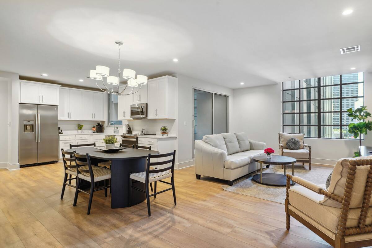 Un appartement moderne comprenant une cuisine, un coin repas et un salon. Il est doté d’une grande fenêtre, d’un lustre et d’une chaise en bois.