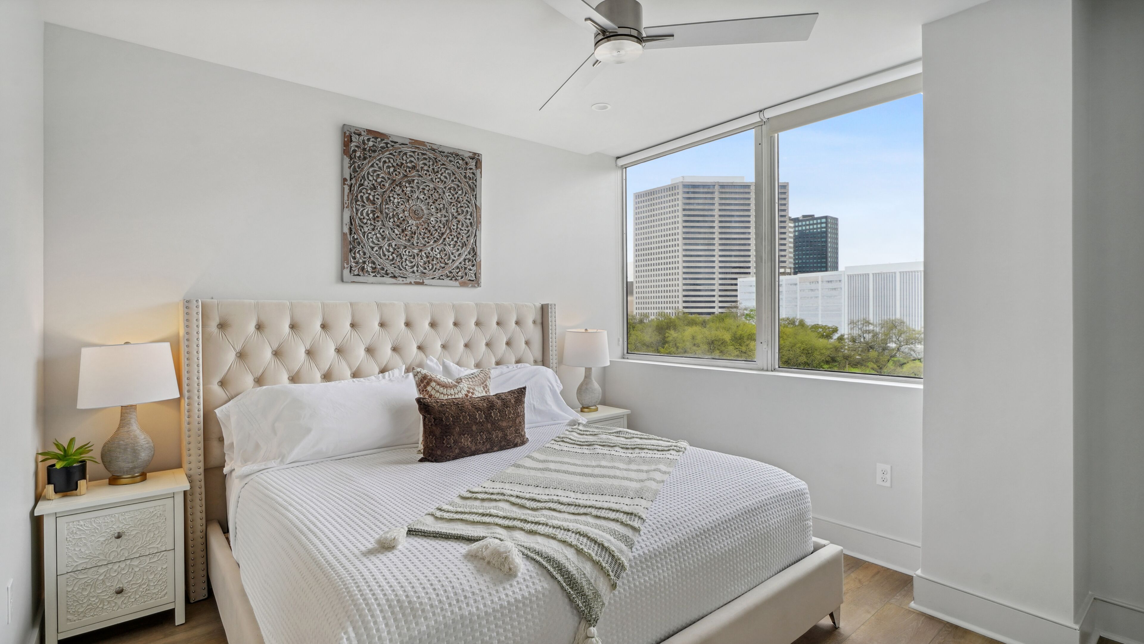 A bedroom with a tufted headboard, a large window with a city view, a decorative wall piece, and a bedside table with a lamp.
