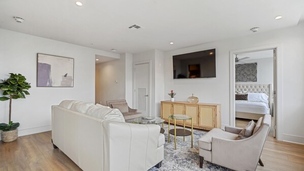 Una sala de estar moderna con un sofá blanco, una mesa de centro de vidrio y un sillón color beige. Se observa un aparador de madera con objetos decorativos y un televisor montado en la pared.