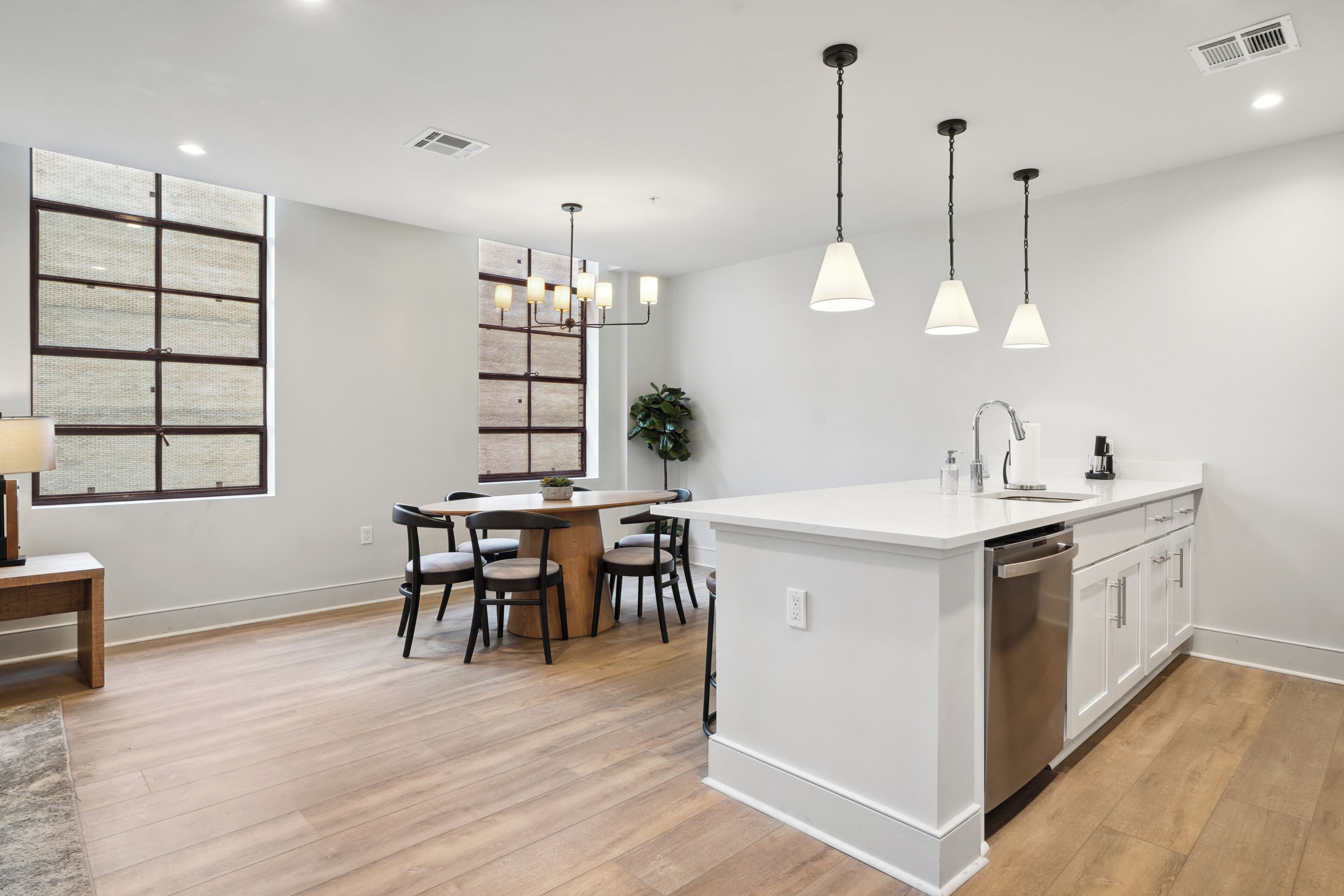 Una cocina moderna con armarios blancos, una isla central, iluminación colgante y una zona de comedor con mesa y sillas de madera.