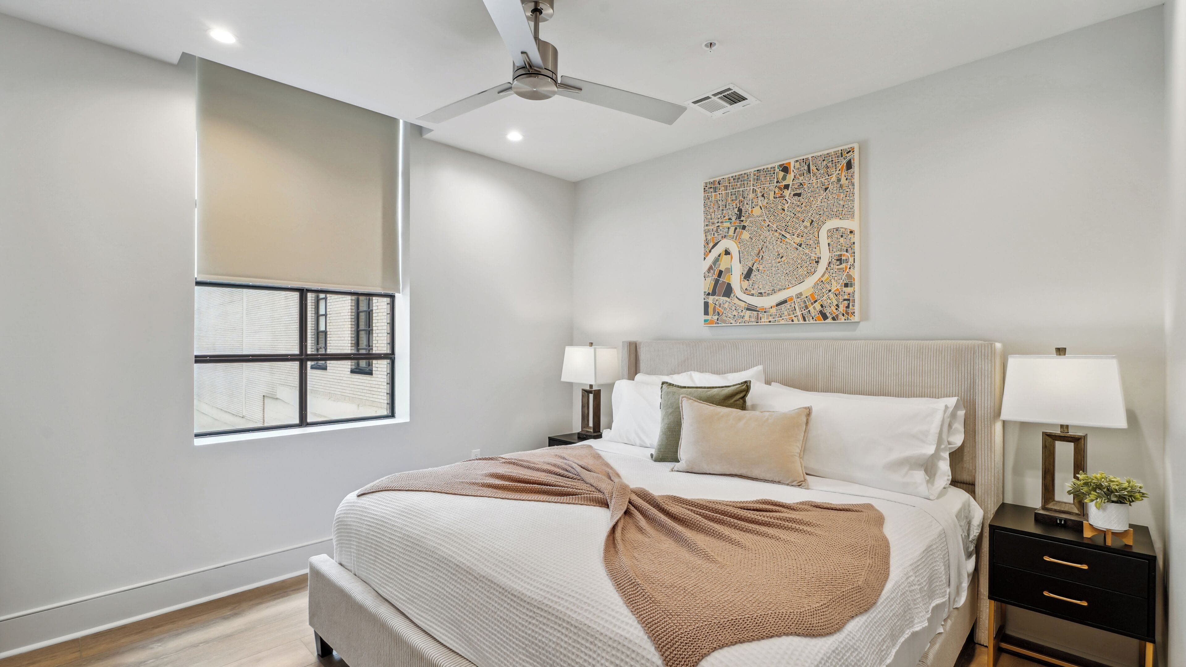 Een moderne slaapkamer met een groot bed, twee nachtkastjes met lampen, een plafondventilator, een raam met uitzicht op gebouwen en een stadsgezichtschilderij aan de muur.