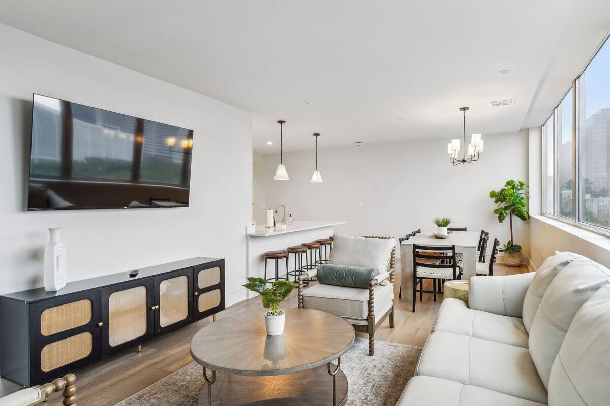 Un salon moderne doté d’un téléviseur à écran plat, d’un coin repas avec des tabourets de bar et d’une grande fenêtre offrant une vue sur la ville.