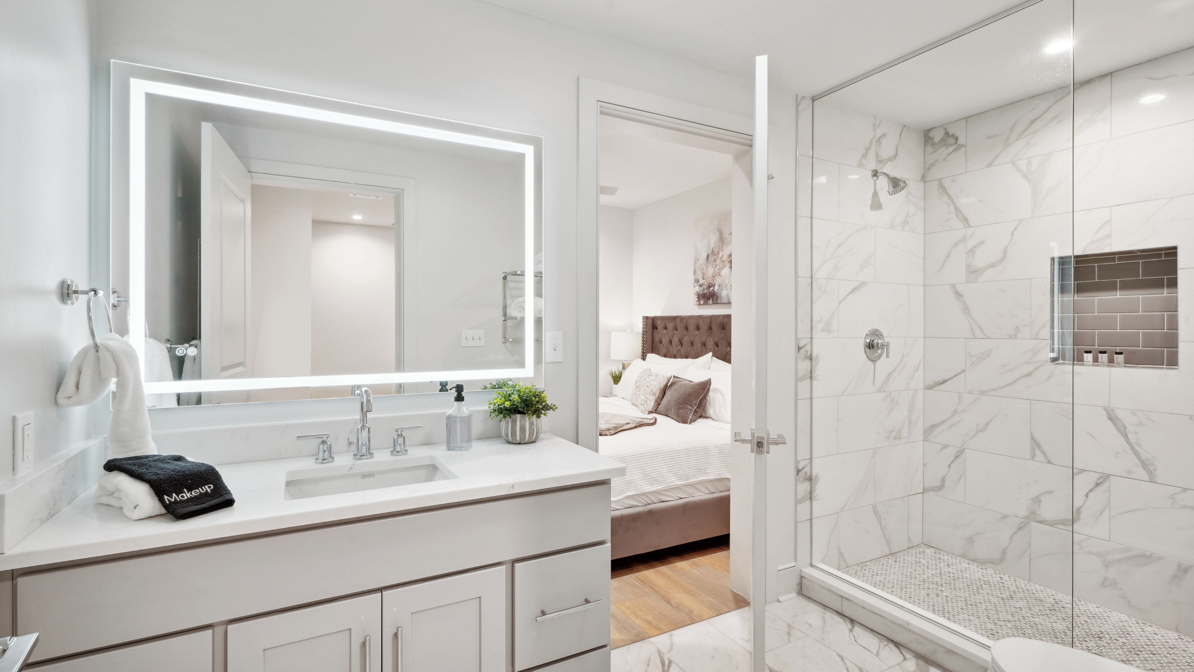 A modern bathroom with a large mirror, a shower with a glass door, and a bedside table with a plant.