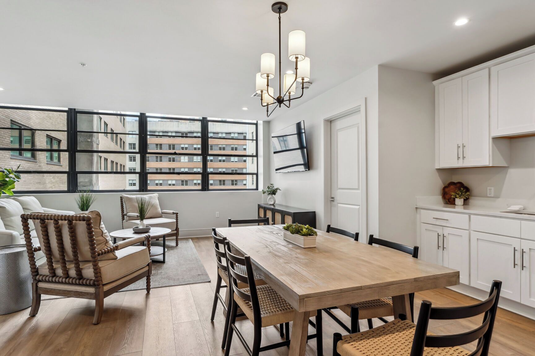 A modern kitchen with a wooden dining table, white cabinets, and a large window overlooking a building.