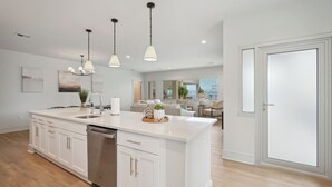 A modern kitchen with a large island, white cabinetry, stainless steel appliances, pendant lighting, and a view to a living area with a balcony.
